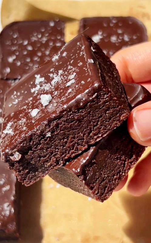 Hand holding no-bake dark chocolate brownie bars with glossy topping and sea salt flakes