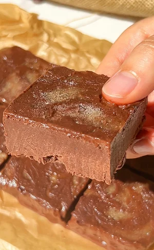 A hand picking up a square of chocolate banana freezer fudge showing the rich gooey fudgy texture on parchment paper