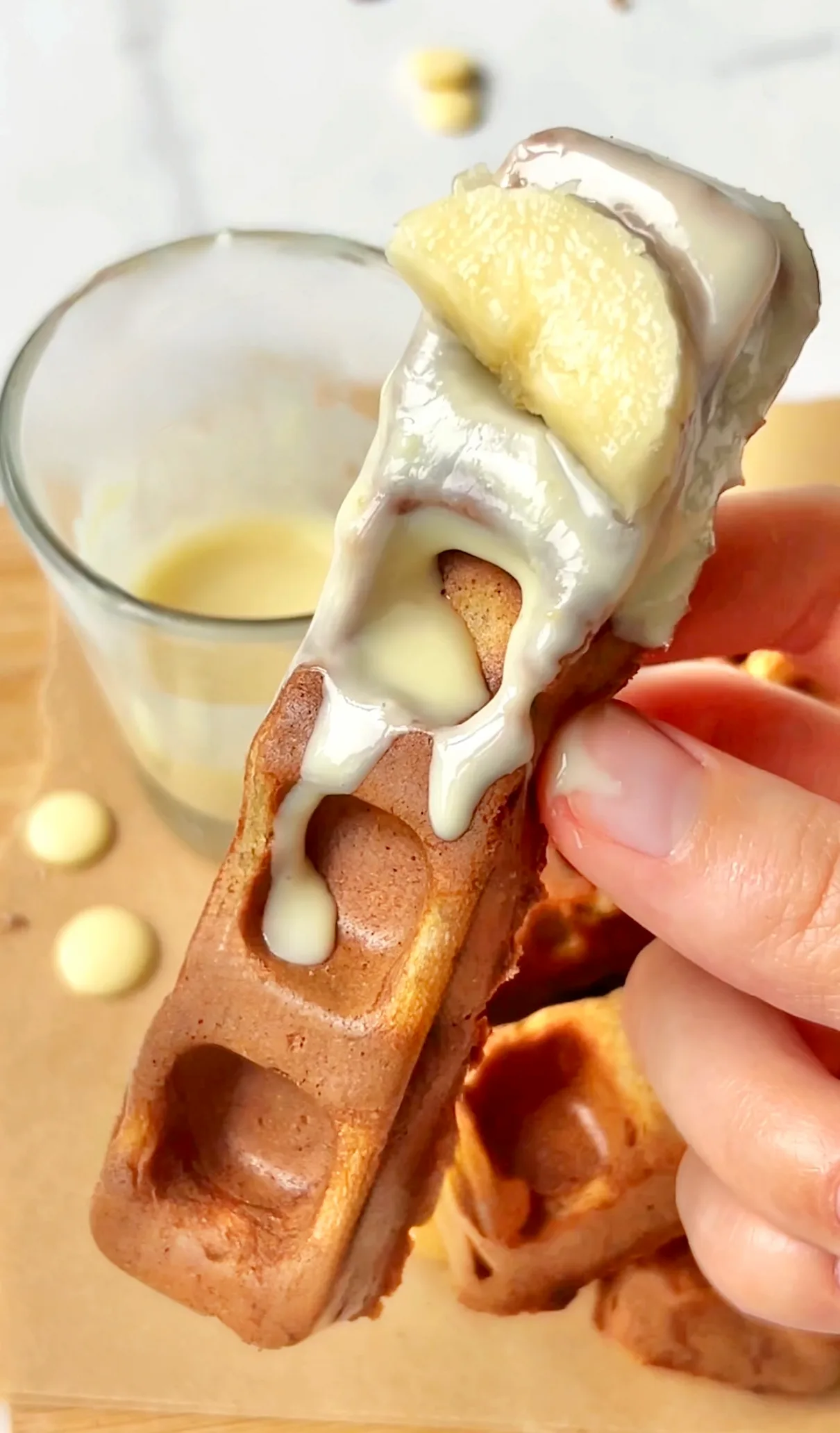 A hand holding a gluten free banana bread waffle stick with white chocolate dripping down and a banana slice on top