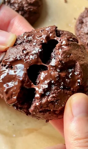 Two hands pulling apart a gluten free brownie cookie revealing the gooey melted chocolate fudgy interior with sea salt flakes on top