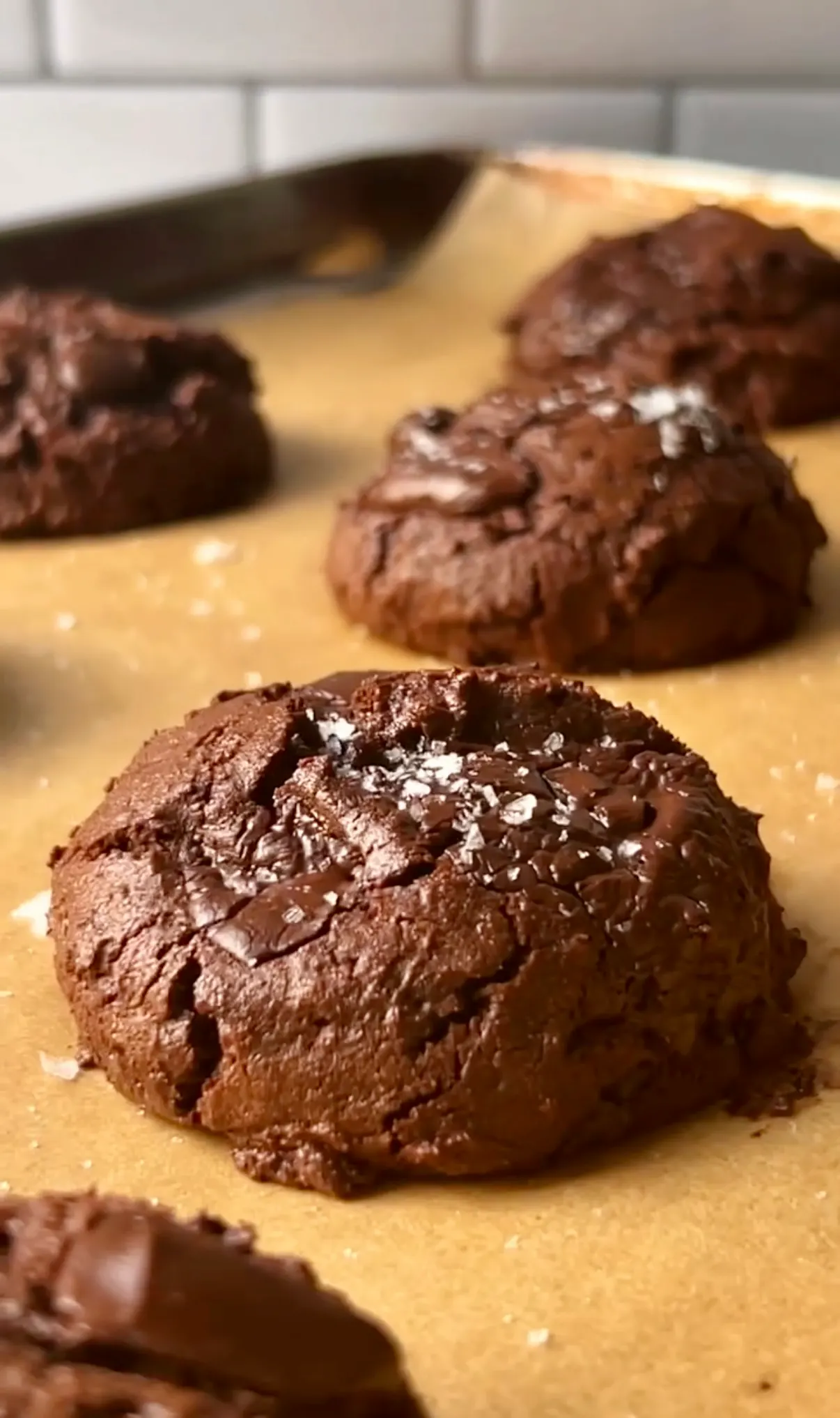 Gluten free brownie cookies on parchment paper on a baking tray showing the crinkled dark chocolate surface with sea salt flakes on top against a white tile background