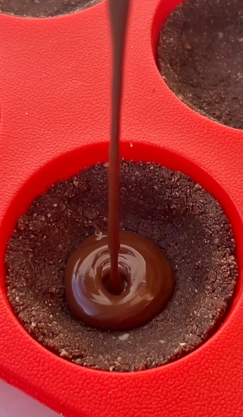 Melted chocolate ganache being poured into a chocolate almond crust cup in a red silicone mould