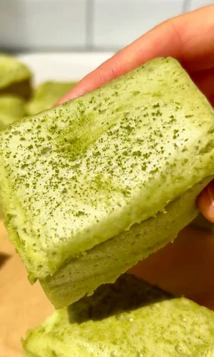 A hand holding a single soft matcha marshmallow square dusted with matcha powder