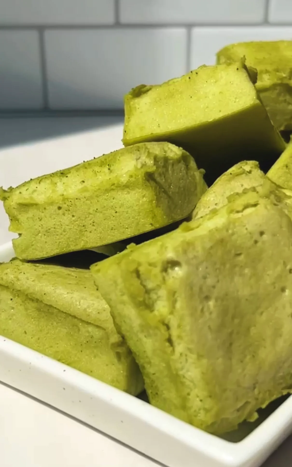 Fluffy homemade matcha marshmallows cut into thick squares and piled in a white dish against a white tile background